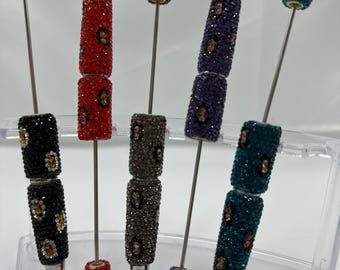 May include: A collection of decorative pens with jewel-encrusted barrels in various colors, including black, red, gray, purple, and teal. Each pen features a silver tip and a matching jewel-topped end. The pens are displayed in a clear plastic stand.