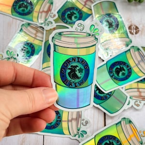 May include: A collection of iridescent holographic stickers featuring a cartoon coffee cup with the text "Morning Brew" and a bird graphic. The stickers are on a light wood surface.