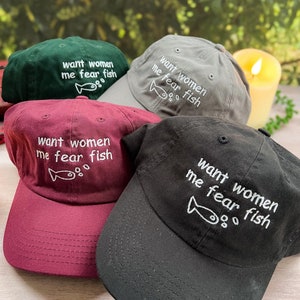May include: Four baseball caps with the text "want women me fear fish" and a small fish drawing embroidered on the front. The caps are in green, grey, burgundy, and black.