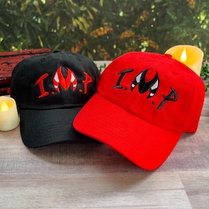 May include: Two baseball caps, one black and one red, with the word "IMP" embroidered in white on the front. The "M" in "IMP" is a stylized image of a devil's horns.