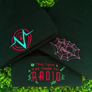 May include: Three black knit beanies with different embroidered designs. The first beanie has a red and blue graphic of a heart with a circle around it. The second beanie has a pink spiderweb with the word "Poison" written inside. The third beanie has the text "This face was made for RADIO" in red and green.