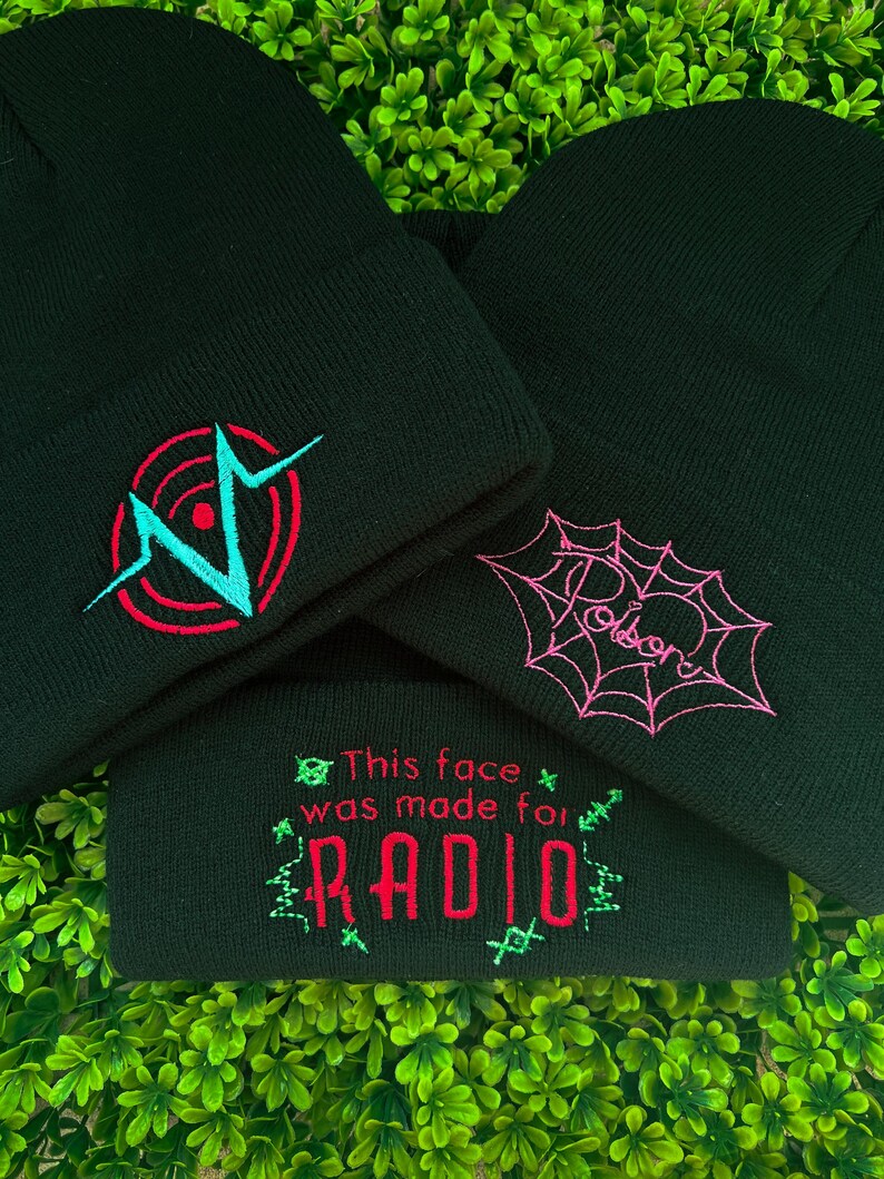 May include: Three black knit beanies with different embroidered designs. The first beanie has a red and blue graphic of a heart with a circle around it. The second beanie has a pink spiderweb with the word "Poison" written inside. The third beanie has the text "This face was made for RADIO" in red and green.