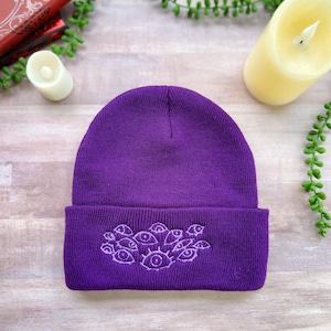 May include: A vibrant purple knit beanie featuring a cuffed brim. The front of the cuff is decorated with an embroidered design consisting of multiple stylized eyes in a lighter shade of purple thread, arranged in a cluster pattern.