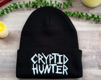 May include: Black knit beanie with white embroidered text that reads "CRYPTID HUNTER".