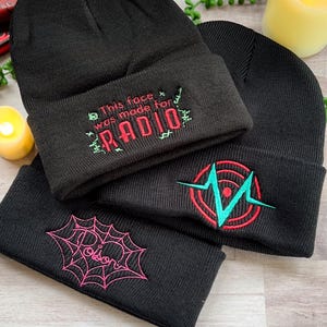 May include: Three black knit beanies with different embroidered designs. The first beanie has the text "This face was made for RADIO" in red and green thread. The second beanie has a red and green heart with a teal lightning bolt inside. The third beanie has a pink spider web with a heart in the center.