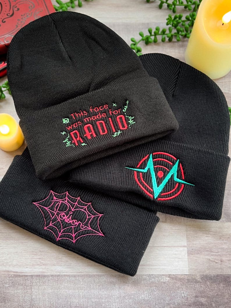 May include: Three black knit beanies with different embroidered designs. The first beanie has the text "This face was made for RADIO" in red and green thread. The second beanie has a red and green heart with a teal lightning bolt inside. The third beanie has a pink spider web with a heart in the center.