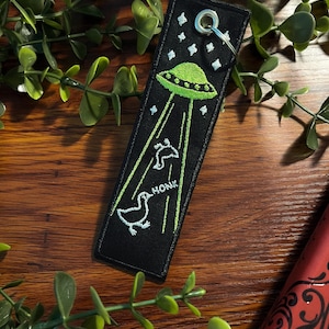 May include: Black fabric keychain with a green UFO and two white ducks. The UFO is emitting beams of light and the ducks are honking. The text "HONK" is embroidered on the keychain.