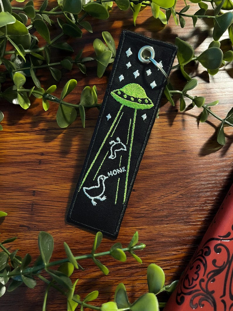 May include: Black fabric keychain with a green UFO and two white ducks. The UFO is emitting beams of light and the ducks are honking. The text "HONK" is embroidered on the keychain.