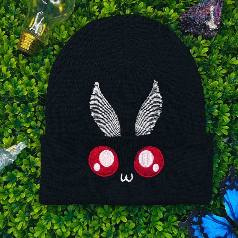 May include: Black beanie with a white embroidered moth design. The moth has red eyes and a cute expression.