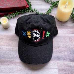 May include: Black baseball cap with a colourful embroidered design. The design includes a cartoon mouth with teeth, dollar signs, exclamation marks, and other symbols. The cap is made of a soft fabric.