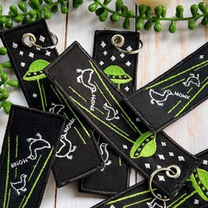 May include: A set of black and green keychains with a UFO and a bird design. The keychains are embroidered with the word "HONK" and have silver key rings.