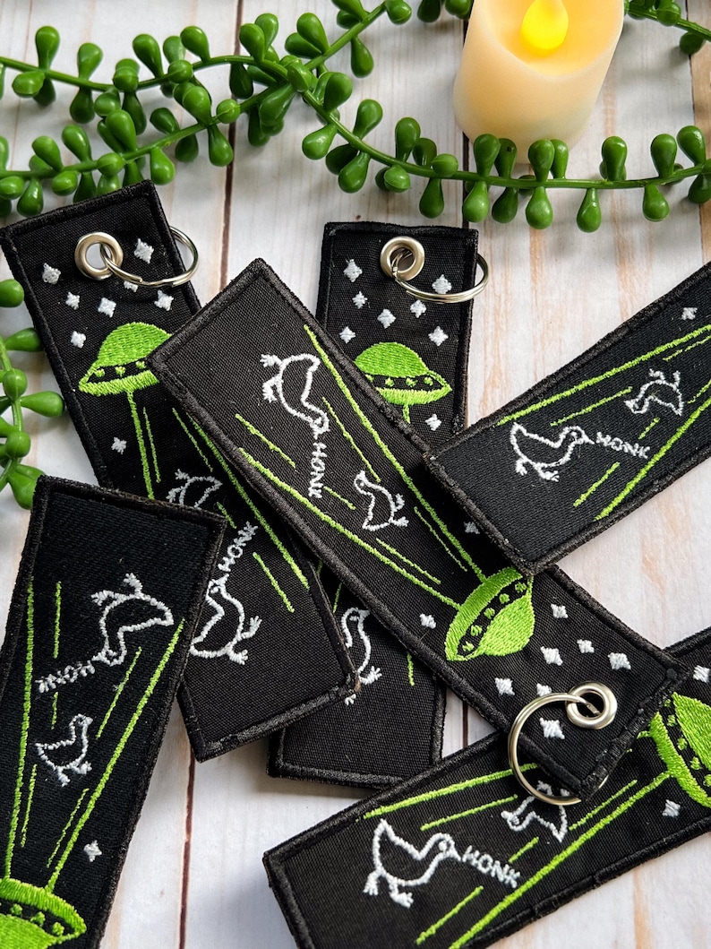 May include: A set of black and green keychains with a UFO and a bird design. The keychains are embroidered with the word "HONK" and have silver key rings.