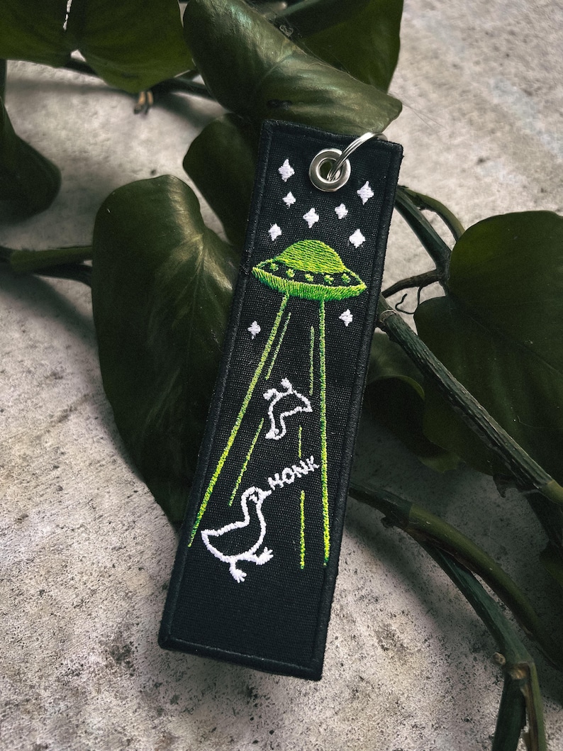 A black fabric keychain with an embroidered image of aliens abducting geese.