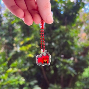 May include: A small, clear acrylic charm featuring a red, cartoon-style creature with sharp yellow teeth and a black bowtie. The charm is attached to a red ball chain, allowing it to hang freely. The design is printed on a transparent material.