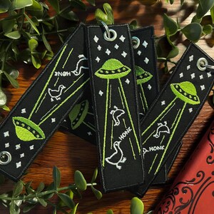 May include: Four black fabric keychains with green and white embroidery. Each keychain features a green UFO with white beams of light and a white goose with the word "HONK" embroidered below. The keychains are arranged in a stack on a wooden surface.