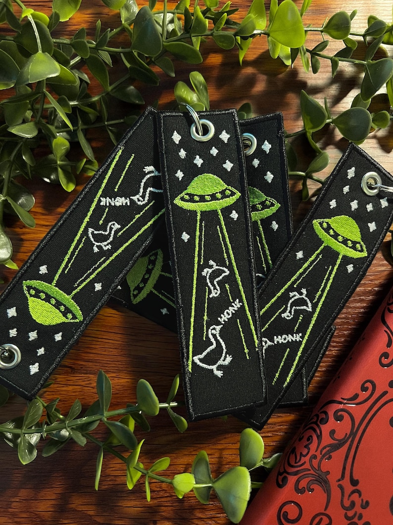 May include: Four black fabric keychains with green and white embroidery. Each keychain features a green UFO with white beams of light and a white goose with the word "HONK" embroidered below. The keychains are arranged in a stack on a wooden surface.