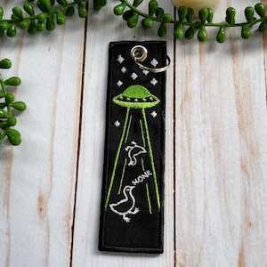 Op de afbeelding: Zwarte sleutelhanger met een groene UFO en drie witte vogels die wegvliegen. De tekst "MOAK" is geborduurd op de sleutelhanger.