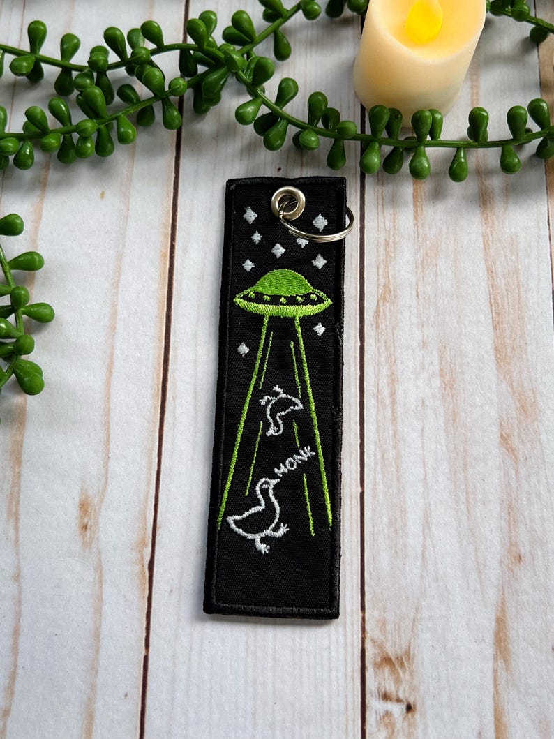 An embroidered jet tag keychain of two geese being abducted by aliens