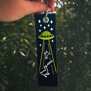 May include: Black key chain with a green UFO and white ducks flying away from it. The key chain has the word "HONK" embroidered on it.