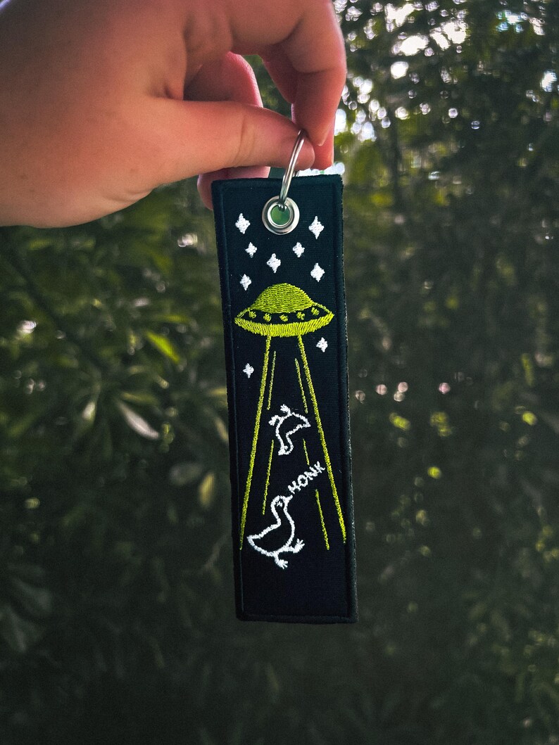 May include: Black key chain with a green UFO and white ducks flying away from it. The key chain has the word "HONK" embroidered on it.