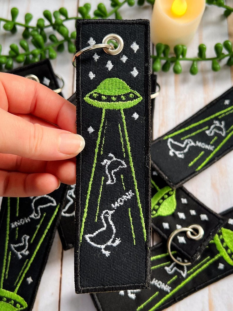 An embroidered jet tag keychain of two geese being abducted by aliens