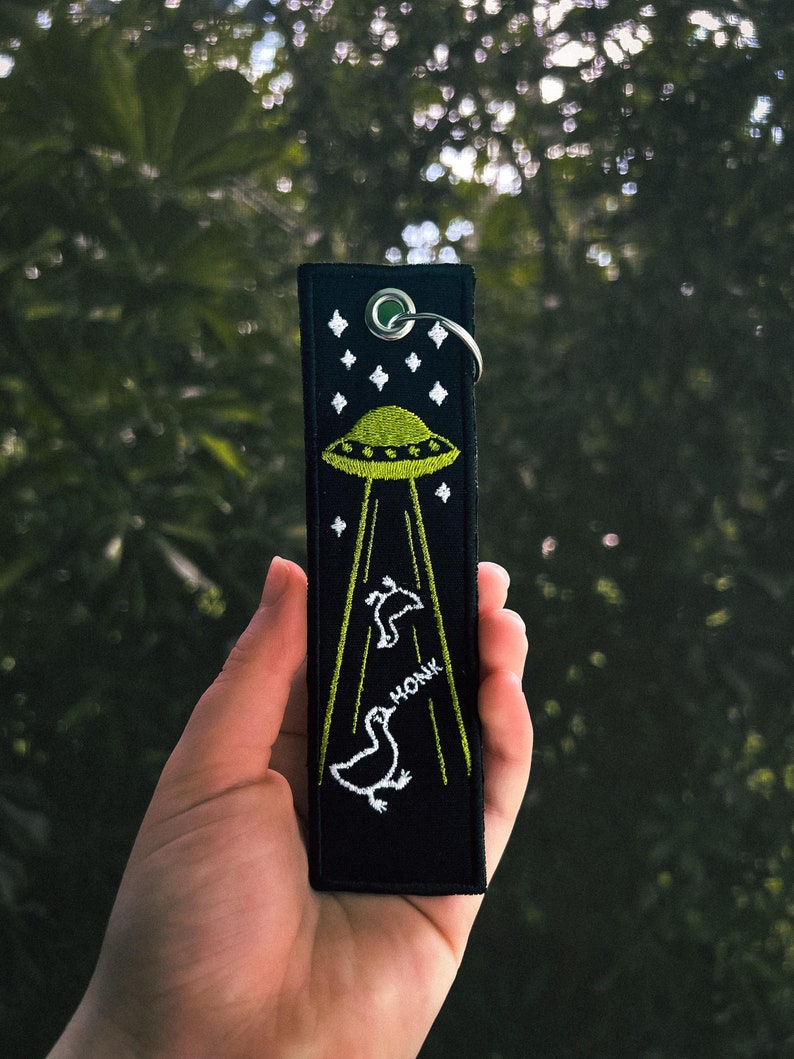 Hand holding a black fabric jet tag keychain with an embroidered image of aliens abducting geese.