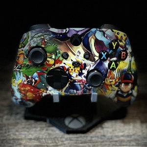 May include: A black Xbox controller with a colorful Pokemon themed design. The controller has a black and white design with a variety of Pokemon characters on it.
