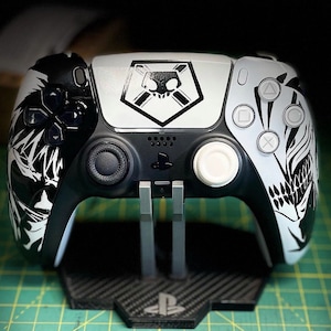 May include: A white and black PlayStation 5 DualSense controller with a skull and crossbones design on the front. The controller is on a black stand with a PlayStation logo.