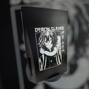 May include: Black and white illustration of a character from the anime Demon Slayer on a black background. The character is wearing a black and white patterned outfit and has a white mask with blue and orange eyes. The text "Demon Slayer" is printed in white above the character. The text "竈門炭治郎" is printed in white to the right of the character.