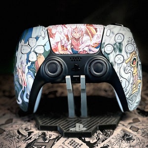 Può includere: Un controller PlayStation 5 bianco e nero con un design anime personalizzato che presenta un personaggio con capelli bianchi e un abito rosa e bianco. Il controller è su un supporto nero con un motivo in fibra di carbonio.