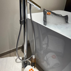 Könnte beinhalten: Eine moderne Badezimmerszene mit einer weißen Badewanne voller Blasen. Eine verchromte Armatur steht hoch, mit einer Handbrause. Weiße Handtücher und eine Seifenflasche sind in der Nähe der Wanne platziert. Ein orange-weißes Rettungsring-Etikett ist an einem Handtuch befestigt.