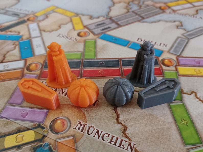 May include: A set of five game pieces for a board game. The pieces are made of plastic and are painted in orange and grey. The pieces include two figures of people wearing cloaks, two pumpkins, and a coffin.