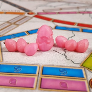 Puede incluir: Cinco piezas de juego de color rosa, una con un dise&ntilde;o de estrella, sobre un tablero de juego con casillas azules y rosas.