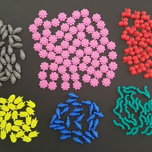 May include: Assortment of colorful plastic game pieces in various shapes, including flowers, fish, leaves, and cherries. The pieces are arranged in groups on a black background.