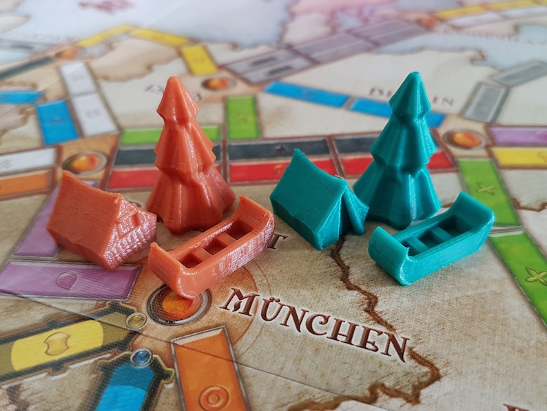 May include: Two sets of game pieces for a board game. One set is orange and includes a house, a tree, and a boat. The other set is teal and includes a tent, a tree, and a boat. The game board is visible in the background with the word "M&Uuml;NCHEN" printed on it.