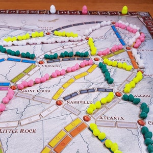 Puede incluir: Un tablero de juego con diferentes piezas de tren de colores en las v&iacute;as. Las v&iacute;as est&aacute;n marcadas con ciudades como Omaha, Kansas City, Saint Louis, Chicago, Nashville, Atlanta, Pittsburgh, Nueva York, Washington, y m&aacute;s. Las piezas de tren son rosas, blancas, amarillas y verdes.