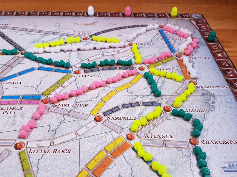 Puede incluir: Un tablero de juego con diferentes piezas de tren de colores en las v&iacute;as. Las v&iacute;as est&aacute;n marcadas con ciudades como Omaha, Kansas City, Saint Louis, Chicago, Nashville, Atlanta, Pittsburgh, Nueva York, Washington, y m&aacute;s. Las piezas de tren son rosas, blancas, amarillas y verdes.