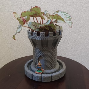 May include: A grey stone tower-shaped planter with a green and white leafy plant growing out of the top. A small toy figure of a person is standing in front of the planter.