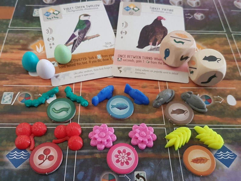 May include: A game board with various colorful game pieces, including birds, eggs, flowers, leaves, and caterpillars. There are also two wooden dice with different animal illustrations. The game board has a light brown background with a white circle in the center.