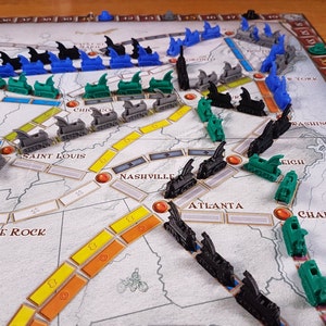 May include: A game board with a map of the United States, featuring trains and cities. The trains are black, green, and blue. The cities are labeled with their names, including Chicago, Saint Louis, Nashville, Atlanta, and Charleston.