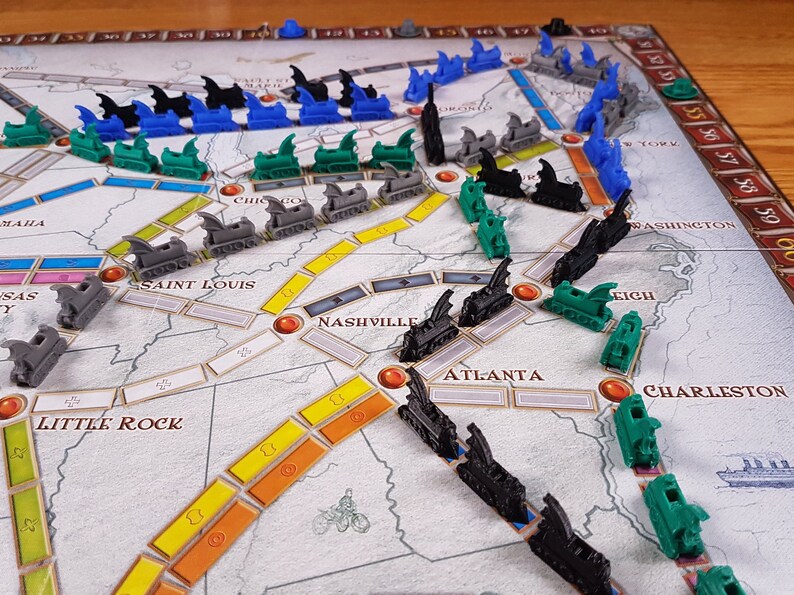 May include: A game board with a map of the United States, featuring trains and cities. The trains are black, green, and blue. The cities are labeled with their names, including Chicago, Saint Louis, Nashville, Atlanta, and Charleston.
