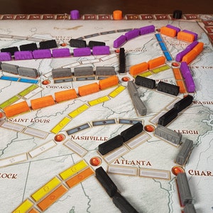 May include: A close-up view of a game board with various colored train pieces. The board features cities across the United States, including Chicago, Saint Louis, Nashville, Atlanta, and New York. The game pieces are arranged in a strategic pattern, suggesting a game in progress.