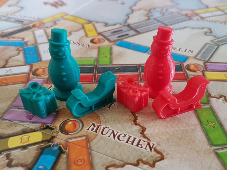 May include: Two snowman game pieces, one green and one red, with matching sleds and presents. The game pieces are on a board game with the word "MUNCHEN" printed on it.