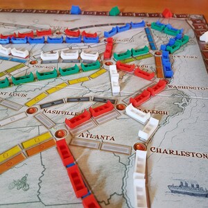 May include: A game board with different colored pieces on it. The board shows a map of the United States with cities labeled. The pieces are red, green, blue, and white.