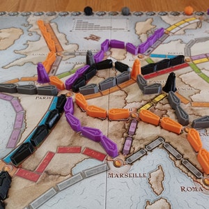 May include: A game board with a map of Europe, featuring train tracks and train pieces in orange, purple, black, and grey. The game board includes city names like Paris, London, and Rome.