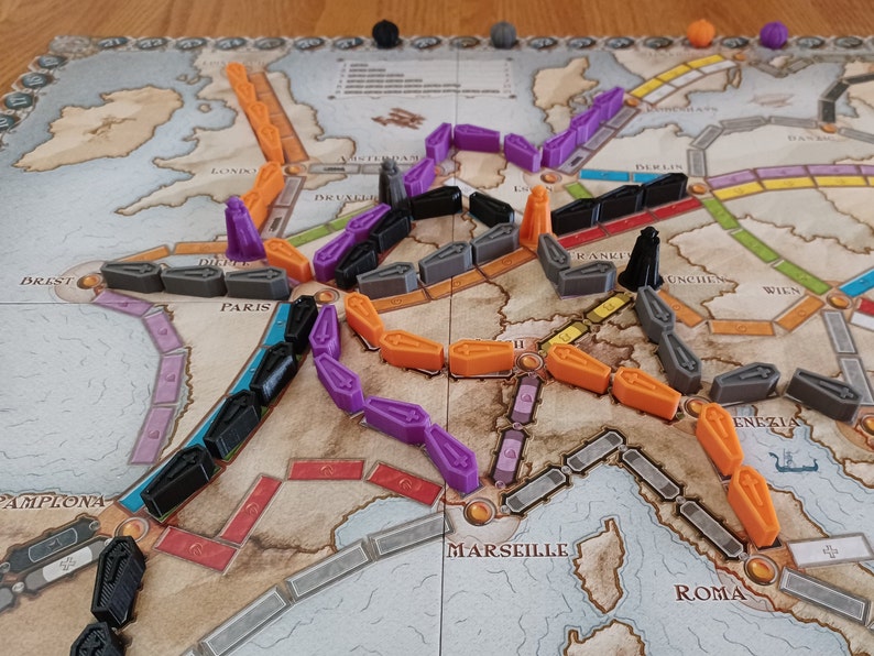 May include: A game board with a map of Europe, featuring train tracks and train pieces in orange, purple, black, and grey. The game board includes city names like Paris, London, and Rome.
