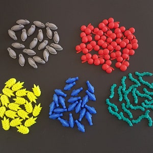 May include: A collection of colorful plastic game pieces in various shapes, including fish, leaves, and berries. The pieces are arranged in groups on a dark gray surface.