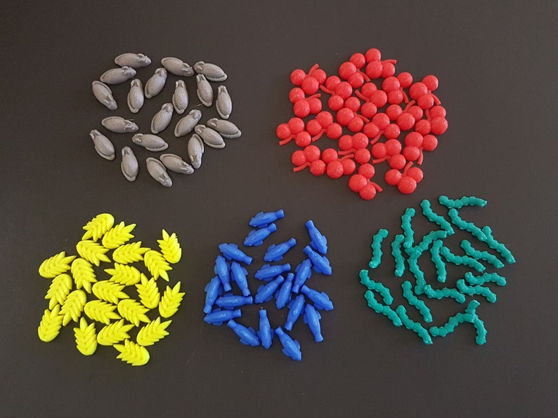 May include: A collection of colorful plastic game pieces in various shapes, including fish, leaves, and berries. The pieces are arranged in groups on a dark gray surface.