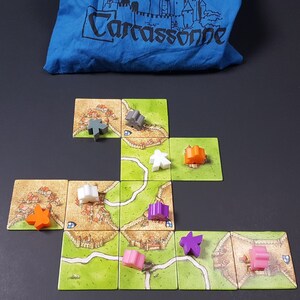 May include: A game board with multiple tiles showing a medieval city with roads and fields. There are several meeples in different colors on the board, including orange, purple, pink, white, and gray.