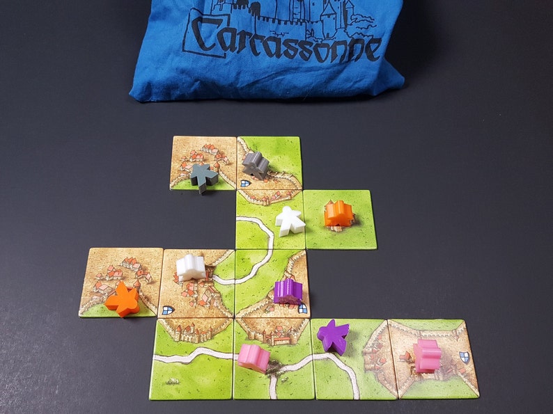 May include: A game board with multiple tiles showing a medieval city with roads and fields. There are several meeples in different colors on the board, including orange, purple, pink, white, and gray.